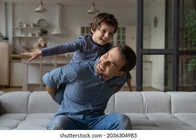 Happy loving daddy and affectionate boy playing active games at home. Dad piggybacking son on couch, kid making airplane wings with open flying hands, laughing, having fun. Family leisure concept - Shutterstock ID 2036304983