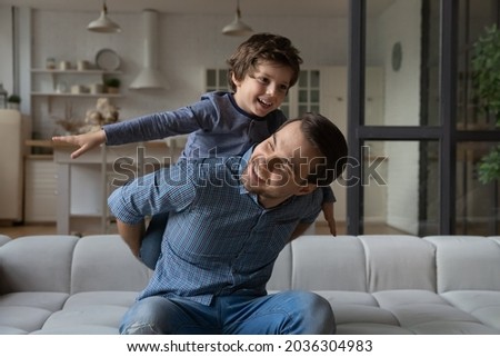 Happy loving daddy and affectionate boy playing active games at home. Dad piggybacking son on couch, kid making airplane wings with open flying hands, laughing, having fun. Family leisure concept