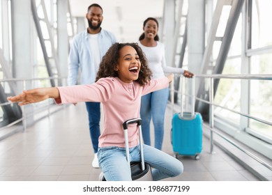 Happy Loving Family In Airport. Portrait of joyful excited black girl sitting on suitcase and laughing, spreading arms imitating plane, smiling cheerful parents walking in blurred background - Powered by Shutterstock