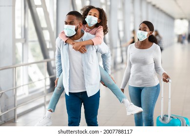 Happy Loving Family In Airport. Portrait Of Black Dad Wearing Face Mask Riding His Daughter On Back, Mom Walking With Suitcase In Terminal Hall. Dad Giving Girl Piggyback Ride, Waiting For Departure