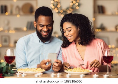 Happy Loving Family. Beautiful Young African American Couple Using Cellphone, Sitting At Table And Having Dinner, Eating Pasta. Smiling Man Sharing Photos With His Woman, Spending Time Together