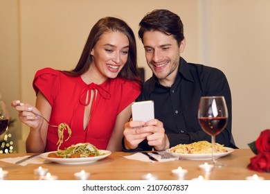 Happy Loving Family. Beautiful Young Millennial Couple Using Cellphone, Sitting At Table And Having Dinner, Eating Pasta. Smiling Man Sharing Photos With His Excited Woman, Spending Time Together