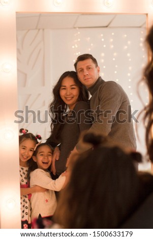 Happy loving family. Cute little girls and and their parents are standing near the mirror in the room in the evening dim light.