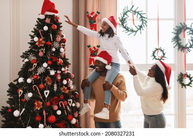 Happy loving family decorating Christmas tree. Young black man holding his daughter on shoulders helping decorate Xmas tree hanging Santa Claus hat on top, people standing in living room together