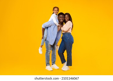 Happy Loving Family. Full Body Length Of Cheerful Black Man, Woman And Girl Standing Posing Isolated On Orange Studio Wall. Cheerful Father Carrying Daughter On Back, Giving Piggyback Ride, Banner