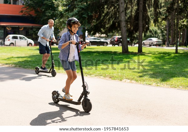 Happy Loving Family. Full body length photo of\
excited curly boy in protective helmet driving motorized push\
scooter with daddy at park, spending time together outdoors at\
summer, selective focus