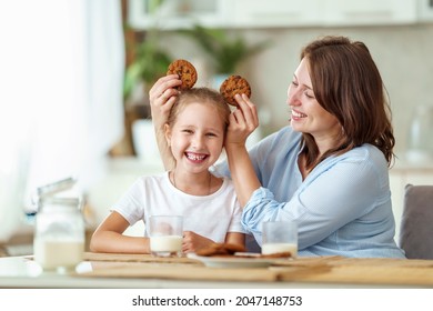 happy loving family, mom and daughter, playing sitting at table and having breakfast at home in morning. woman and girl eat oatmeal cookies and drink cow's milk, and have nice time together in kitchen