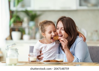 Happy Loving Family, Mom And Daughter, Playing Sitting At Table And Having Breakfast At Home In Morning. Woman And Girl Eat Oatmeal Cookies And Drink Cow's Milk, And Have Nice Time Together In Kitchen