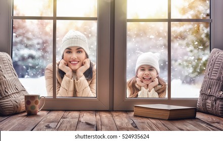 Happy loving family! Mother and child girl having fun, playing and laughing on snowy winter walk in nature. Frost winter season. Child and mom looking in windows, standing outdoors. View indoors home.