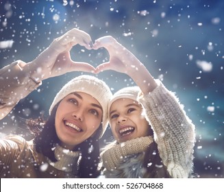 Happy Loving Family! Mother And Child Girl Having Fun, Playing And Laughing On Snowy Winter Walk In Nature. Frost Winter Season. Mom And Kid Making Hand Heart In Evening.