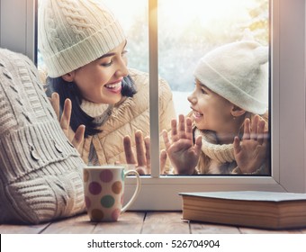 Happy Loving Family! Mother And Child Girl Having Fun, Playing And Laughing On Snowy Winter Walk In Nature. Frost Winter Season. Child And Mom Looking In Windows, Standing Outdoors. View Indoors Home.