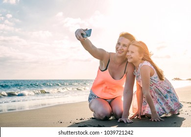 Happy Loving Family Mother And Daughter Taking A Selfie With Mobile Smart Phone On The Beach At Sunset - Mom With Her Kid In Holiday - Parent, Vacation, Tech And Family Lifestyle Concept