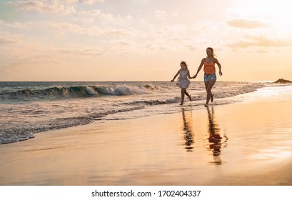 Happy Loving Family Mother And Daughter Running On The Beach At Sunset - Mum Having Fun With Her Kid Long Sea Shore During Summer Holidays - Parent Vacation Time Lifestyle Concept