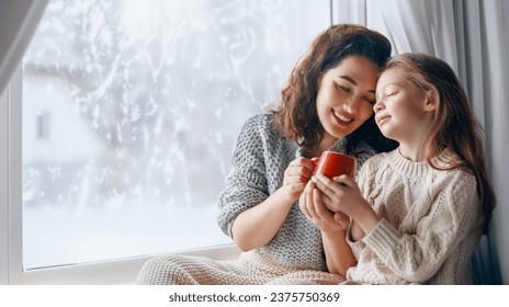 Happy loving family. Mother and daughter are hugging and drinking hot beverage enjoying winter nature in the  window. - Powered by Shutterstock
