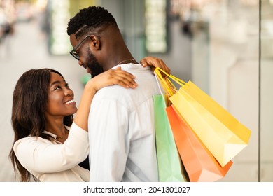 Happy Loving Family. Portrait Of Affectionate Smiling African American Woman Embracing Her Man, Carrying Shopping Bags And Looking At Him Walking Near Mall, Black Couple Hugging, Blurred Background