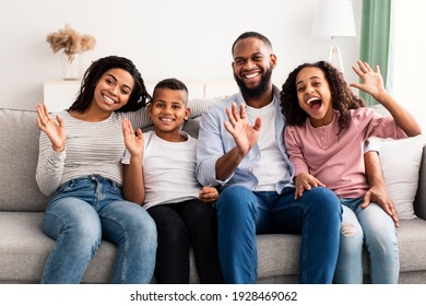 Happy Loving Family. Portrait Of Cheerful African American Man, Woman, Boy And Girl Sitting On The Sofa At Home, Posing For Photo And Waving Hands To Camera. Four Smiling Young Black People