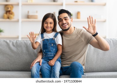 Happy Loving Family. Portrait Of Cheerful People Posing For Photo And Waving Hands To Camera, Young Girl Sitting On Father's Lap, Man With His Daughter Resting On The Sofa At Home