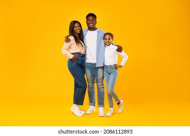 Happy Loving Family. Portrait Of Three African American People Standing Isolated Over Yellow Orange Studio Wall, Posing At Camera. Smiling Husband Hugging His Wife And Daughter. Togetherness Concept