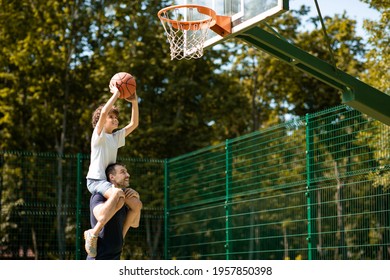 Happy Loving Family. Portrait Of Young Curly Boy Sitting On Father's Shoulders, Playing Basketball, Throwing Ball To Basket, Man Standing On Court Outdoors And Helping His Son, Selective Focus