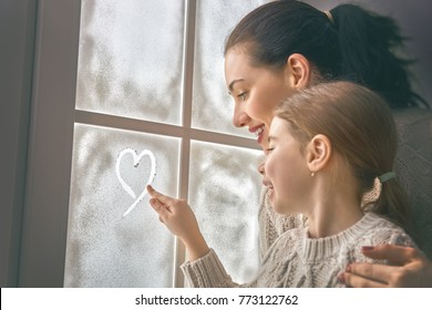 Happy loving family sitting by the window and drawing a heart on frozen glass. Mother and child creating decorations. Concept of love and care.