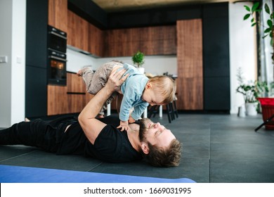 Happy Loving Father Lying On A Floor Of An Apartment, Lifting His Baby In The Air. Smiling, Laughing.