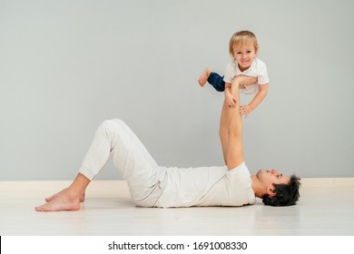 Happy Loving Father In Sportswear Lying On A Floor Of Grey Wall Background, Doing Press Exercise, Playing With His Son Lifting His Baby In The Air. Smiling, Laughing, Relaxing, Man Solidarity Concept.