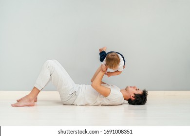 Happy Loving Father In Sportswear Lying On A Floor Of Grey Wall Background, Doing Press Exercise, Playing With His Son Lifting His Baby In The Air. Smiling, Laughing, Relaxing, Man Solidarity Concept.