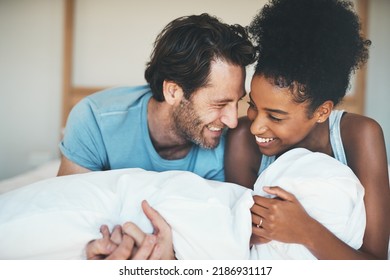 Happy, Loving And Funny Interracial Couple Smiling And Laughing While Lying In Bed And Sharing A Tender Moment. Carefree Husband And Wife Relaxing And Having Fun While Bonding In The Bedroom