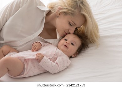 Happy loving mom kissing sweet few month baby with care, affection, tenderness, resting on bed mattress with white sheet. New mother enjoying leisure time with little infant kid in bedroom