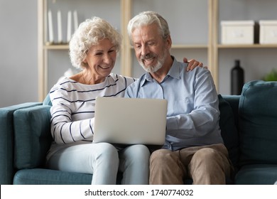 Happy Loving Older Family Spouses Sitting On Sofa, Looking At Computer Screen. Smiling Middle Aged Man Showing Funny Video On Laptop To Excited Mature Wife, Retirement Weekend Lifestyle Concept.