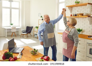 Happy Loving Senior Couple In Aprons Dancing And Having Fun While Cooking Healthy Meal In The Kitchen Of Their Studio Apartment. Seniors Leading Active Lifestyle, Enjoying Life, Spending Time Together