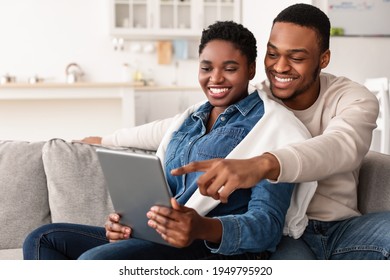 Happy Loving Young African American Couple Using Digital Tablet, Watching Video, Looking At Their Photos Or Choosing What To Buy, Sitting And Relaxing On Couch In Living Room. Man Pointing At Screen