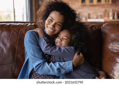 Happy Loving Young African American Mother And Small Teen Daughter Relax On Sofa Hug Cuddle At Home Together. Smiling Caring Ethnic Mom And Little Biracial Girl Child Embrace. Motherhood Concept.