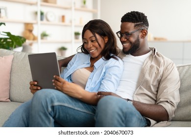 Happy Loving Young African American Couple Using Digital Tablet, Watching Video, Looking At Their Photos Or Choosing What To Buy, Sitting And Relaxing On Couch In Living Room. Guy Embracing Lady