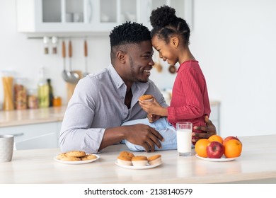 Happy Loving Young Black Father Hugging Smiling Little Daughter, Bonding At Kitchen While Having Snack Together, Girl Sitting On Table, Holding Cupcake, Drinking Milk, Copy Space. Fatherhood Concept