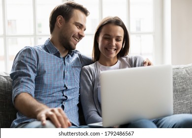 Happy Loving Young Man Cuddling Smiling Pretty Wife, Watching Comedian Movie Or Funny TV Series On Laptop. Emotional Joyful Bonding Couple Looking At Computer Screen, Web Surfing Or Shopping Online.