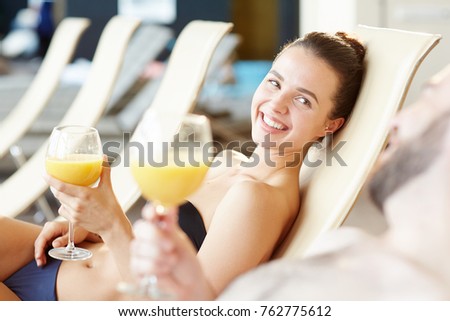 Happy luxurious female with glass of juice talking to man while relaxing on deckchair at spa resort