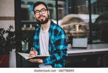 Happy Male Blogger In Optical Eyewear For Vision Correction Pondering On Idea For University Essay, Asian Hipster Student Holding Textbook For Education During Time For Autodidact In Street Cafeteria