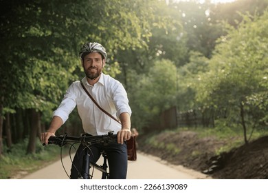 Happy male businessman in protective helmet, cycling along park alley outdoors. Front view of of young man in shirt looking at camera, smiling, copy space. Eco friendly concept. - Powered by Shutterstock