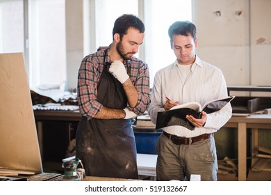 Happy male carpenter showing something to coworker at his notebook papers in workshop - Shutterstock ID 519132667