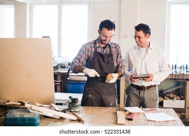 Happy male carpenter showing something to coworker at his notebook papers in workshop