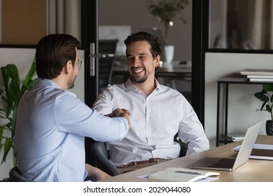 Happy male colleagues employees shake hands working together at laptop at team meeting in office. Smiling business partners or clients handshake close deal make agreement. Employment concept. - Shutterstock ID 2029044080
