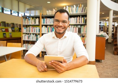 Happy male customer using public Wi-Fi hotspot in library. Young Latin man sitting at desk, smiling at camera and holding tablet. Wireless internet connection concept - Powered by Shutterstock