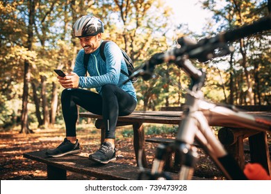 Happy male cyclist sitting in forest and texting message on his mobile phone. Soft focus image. - Powered by Shutterstock