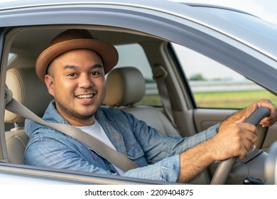 Happy Male Driver Smiling While Sitting In A Car With Open Front Window. Young Asian Man Smile And Looking Through Window. View Of A Young Man Driving His Car To Travel On His Holiday Vacation Time.