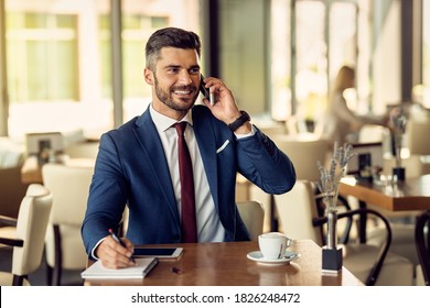 Happy male entrepreneur writing in notebook while talking on cell phone during his coffee break in a cafe.  - Powered by Shutterstock