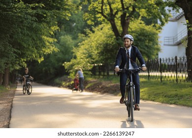 Happy male executive in business outfit returning from job on bicycle outdoor. Front view of determined office clerk feeling great, while cycling home from office in city park. Concept of transport. - Shutterstock ID 2266139247