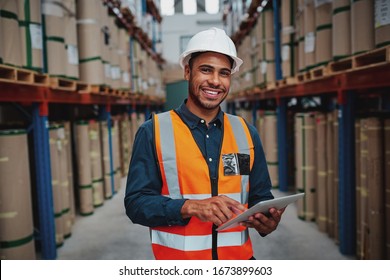 Happy Male Factory Manager Using Digital Tablet In Warehouse While Standing Against Goods Shelf Looking At Camera