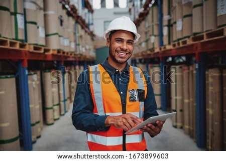 Happy male factory manager using digital tablet in warehouse while standing against goods shelf looking at camera