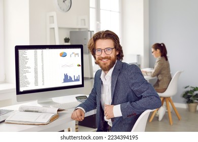 Happy male financial accountant data analyst at work in office  Handsome bearded young man in suit   eyeglasses sitting at computer screen and graphs   charts  looking at camera   smiling